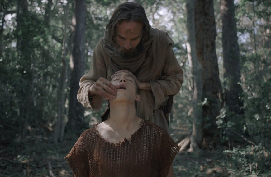 A long-haired man in linen feeds a sacrament to a kneeling young man