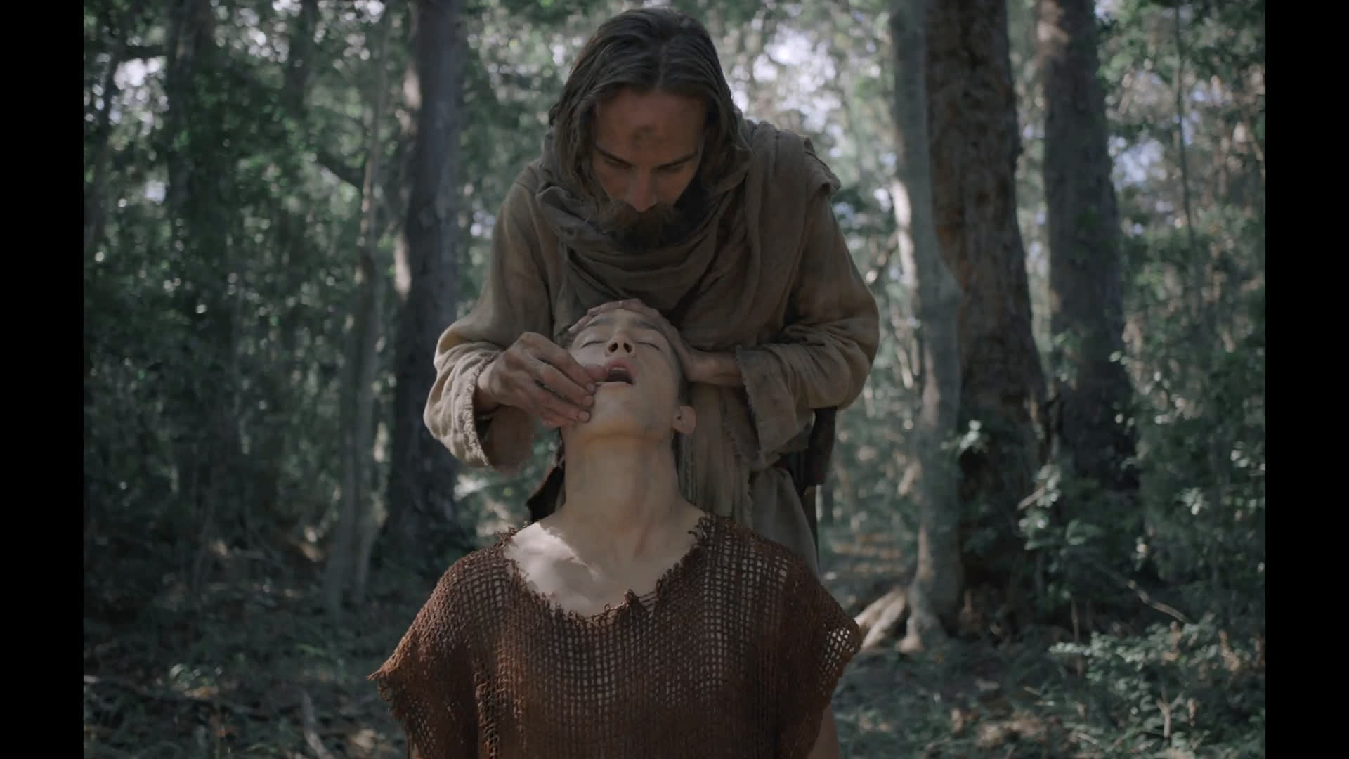 A long-haired man in linen feeds a sacrament to a kneeling young man