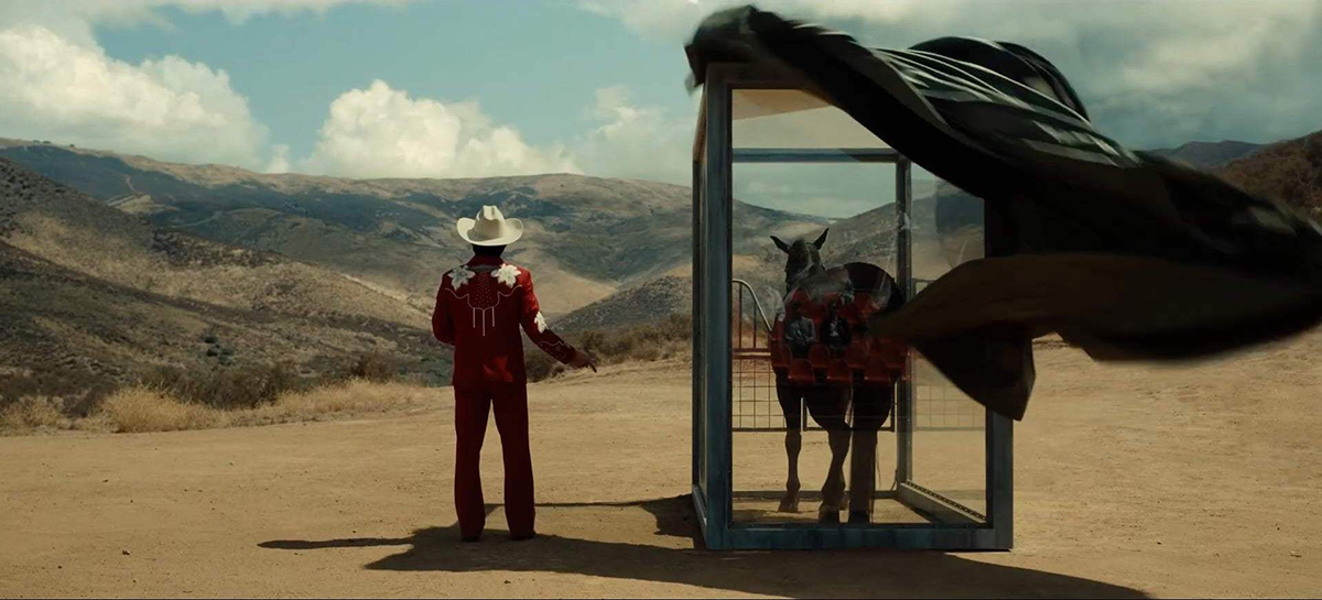 A still from the film Nope showing a man in a red cowboy suit and white cowboy hat next to a horse inside a glass box